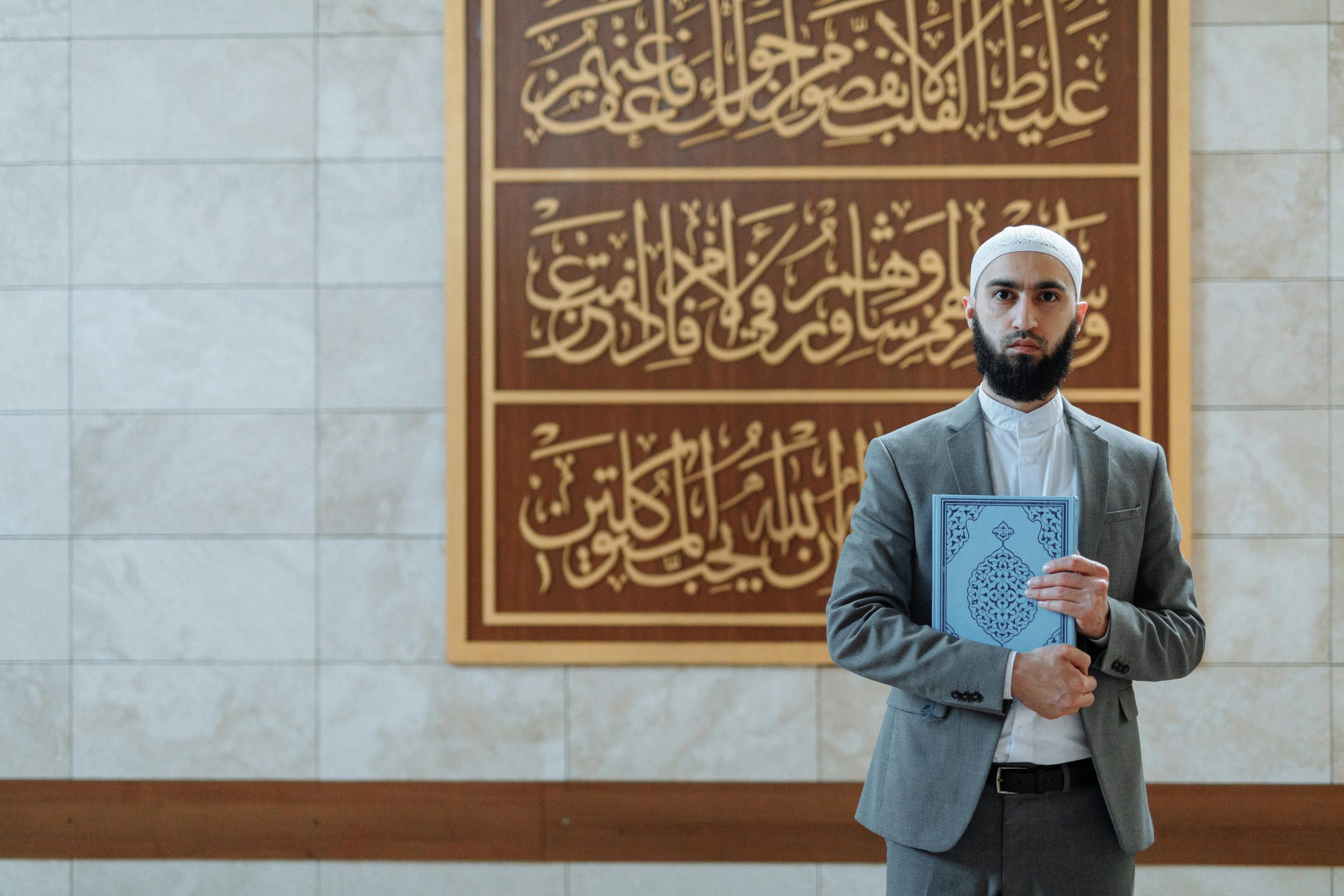 A bearded Muslim man in traditional attire holds a Quran inside a mosque, symbolizing faith and spirituality.